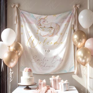 Pode incluir: Uma cena de ch&aacute; de beb&eacute; com uma faixa com uma ilustra&ccedil;&atilde;o de unic&oacute;rnio e as palavras "Welcome to Emilia Baby Shower". A mesa est&aacute; decorada com um bolo, presentes e bal&otilde;es dourados, brancos e rosa.