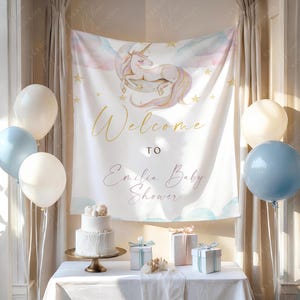 May include: A baby shower setup with a white tablecloth, balloons, and a banner reading "Welcome to Emilia Baby Shower." A two-tiered cake is on a gold stand, with gifts on the table. The banner displays a unicorn illustration.