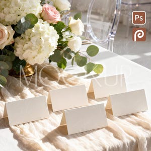 May include: A table setting with a floral arrangement of white hydrangeas, roses, and eucalyptus. Six blank, folded place cards are arranged on a cream-colored fabric. A clear chair is in the background.