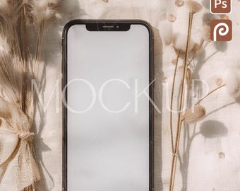 May include: A smartphone with a blank white screen, framed in black, rests on a beige linen surface. Dried beige ornamental grasses and small decorative elements surround the phone. The word "MOCKUP" is faintly visible on the screen.