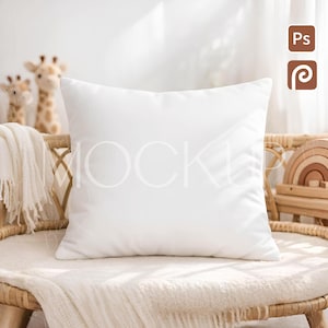 May include: A square, white pillow rests on a wicker chair draped with a cream-colored blanket. Two giraffe stuffed animals and a wooden rainbow toy are visible in the background. The pillow is the focal point, suggesting a home decor theme.