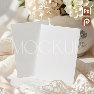 Puede incluir: Dos tarjetas blancas en blanco se muestran sobre una superficie blanca, con la palabra "MOCKUP" visible. Un ramo de flores blancas se encuentra en un jarrón blanco en el fondo. La escena está decorada con encaje y una cinta.