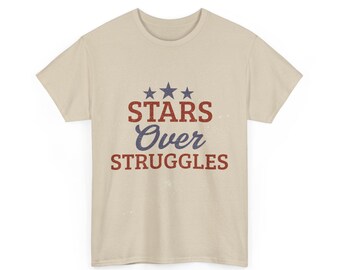 Può includere: Una maglietta beige con la scritta "STARS Over STRUGGLES" in rosso e blu. Il design include tre stelle blu sopra la parola "STARS". La maglietta ha un girocollo classico e maniche corte.