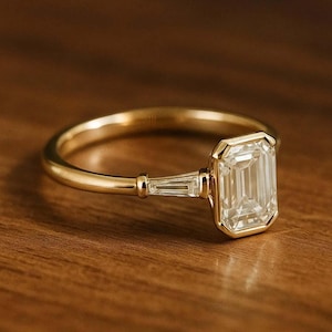 May include: A gold ring with an emerald-cut diamond center stone and two baguette diamonds on the sides. The ring is set on a thin gold band and is displayed on a wooden surface.