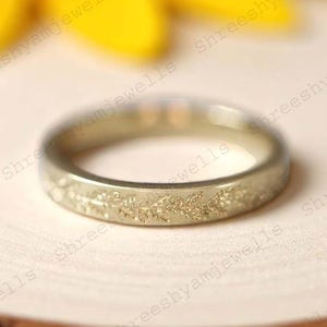 Może przedstawiać: Złoty pierścionek z kwiatowym wzorem wytrawionym wokół obrączki. Pierścionek jest osadzony na drewnianej powierzchni, z rozmytym żółtym kwiatem w tle. Pierścionek jest w prostym stylu obrączki.