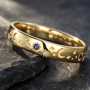 Puede incluir: Un anillo dorado con diseños celestiales. El anillo presenta medias lunas, estrellas y soles grabados, con pequeñas piedras preciosas azules y claras. El anillo está sobre un fondo oscuro y texturizado.