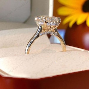 Pode incluir: Um anel de noivado de ouro com uma grande pedra central transparente de corte oval. O anel apresenta um halo de pedras menores ao redor da pedra central e pedras adicionais na banda. O anel é cravejado em uma banda de ouro.