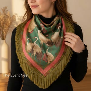 Peut inclure: Un foulard carré avec un fond vert orné de grues blanches et de motifs floraux rouges. Il présente une large bordure rose à motifs décoratifs et est agrémenté de longues franges vert olive sur les bords. The Event Nest.