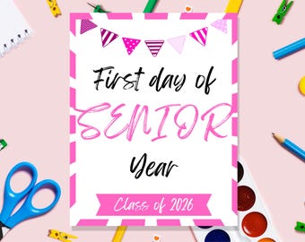 May include: A pink and white sign with the text "First day of SENIOR Year" and "Class of 2026". The sign has a pink banner design at the top and bottom. The background is a light pink surface with school supplies.