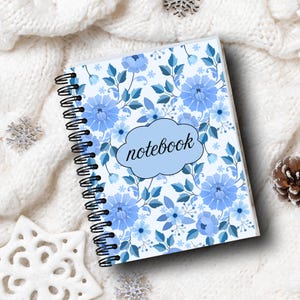 May include: A spiral-bound notebook with a floral design featuring blue flowers and leaves. The cover has the word "notebook" in a light blue cloud-shaped text box. The notebook is on a white, textured surface with decorative snowflakes.