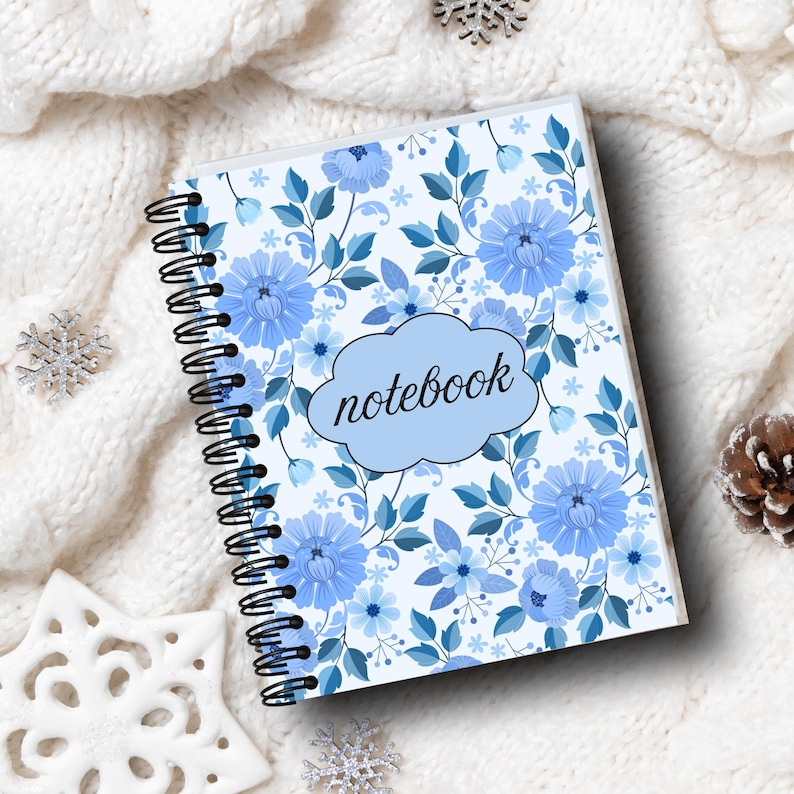 May include: A spiral-bound notebook with a floral design featuring blue flowers and leaves. The cover has the word "notebook" in a light blue cloud-shaped text box. The notebook is on a white, textured surface with decorative snowflakes.