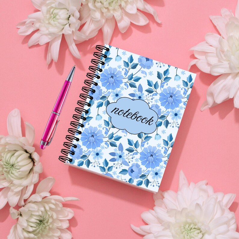May include: A spiral-bound notebook with a blue floral pattern and the word "notebook" on the cover. A pink pen rests beside the notebook. White flowers are arranged around the notebook on a pink background.