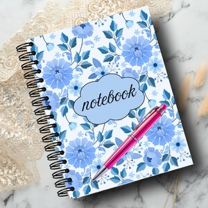 May include: A spiral-bound notebook with a floral design in shades of blue and white. The word "notebook" is in a light blue cloud-shaped text box. A pink pen rests on the notebook. The notebook is on a marble surface.