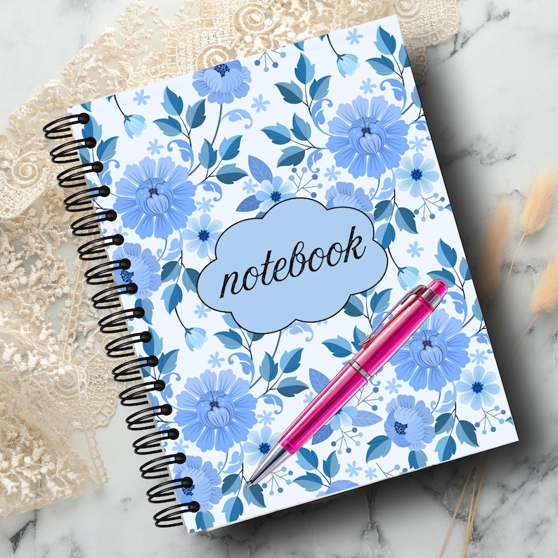 May include: A spiral-bound notebook with a floral design in shades of blue and white. The word "notebook" is in a light blue cloud-shaped text box. A pink pen rests on the notebook. The notebook is on a marble surface.