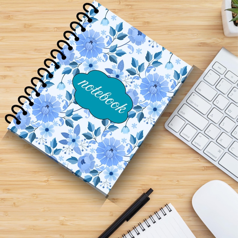 May include: A spiral-bound notebook with a floral design featuring blue flowers and leaves on a white background. The word "notebook" is written in teal on a teal cloud-shaped label. A keyboard, mouse, pen, and small notepad are also visible.
