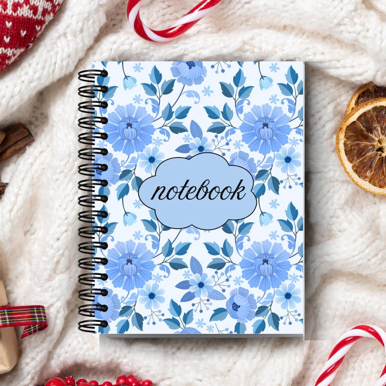 May include: A spiral-bound notebook with a floral pattern in shades of blue. The cover features a light blue cloud-shaped text box with the word "notebook" in black script. The background is a white knit fabric.
