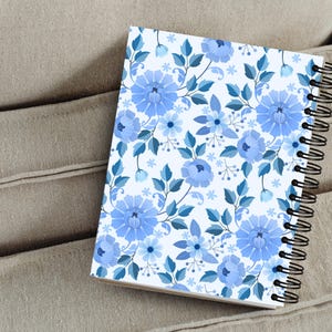 May include: A spiral-bound notebook featuring a floral pattern in shades of blue. The design includes large, stylized blue flowers, smaller blossoms, and leaves on a white background. The notebook's black spiral binding is visible along the edge.