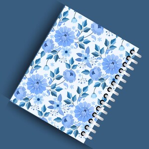May include: A spiral-bound notebook with a floral pattern. The cover features a repeating design of blue flowers and leaves on a white background. The notebook's spiral binding is white, and the overall aesthetic is soft and feminine.