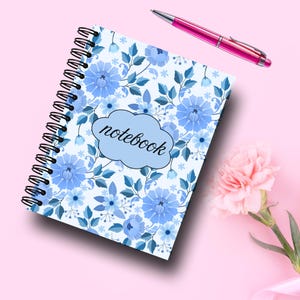 May include: Spiral-bound notebook with a blue floral pattern and the word "notebook" on the cover. A pink pen rests beside the notebook on a pink surface. A pink flower is in the lower right corner.