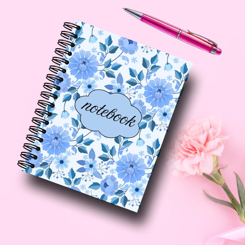 May include: Spiral-bound notebook with a blue floral pattern and the word "notebook" on the cover. A pink pen rests beside the notebook on a pink surface. A pink flower is in the lower right corner.