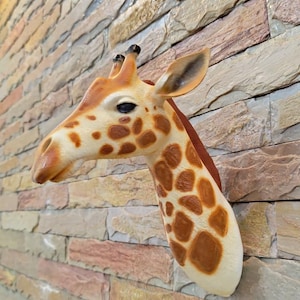 May include: A giraffe head sculpture with a cream-colored face, brown spots, and a reddish-brown neck. The sculpture is mounted on a textured stone wall, creating a striking contrast. The giraffe's eyes are dark, and its ears are detailed.