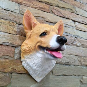 Puede incluir: Escultura de cabeza de perro corgi montada en la pared. La escultura presenta un diseño realista con pelaje marrón y blanco, nariz negra y lengua rosa. La cabeza del perro está montada en una pared de piedra.