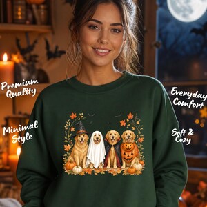 Könnte beinhalten: Ein dunkelgrünes Sweatshirt mit Rundhalsausschnitt und einem Grafikdruck von vier Golden Retrievern in Halloween-Kostümen, darunter ein Hexenhut, ein Geisterlaken, ein Vampirumhang und ein Kürbiskostüm, umgeben von Herbstblättern und Kürbissen.