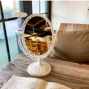 Peut inclure: Un miroir de courtoisie ovale blanc avec des détails ornés. Le miroir mesure 28 cm x 19 cm et repose sur une base décorative. Le cadre et la base présentent un motif de volutes. Le miroir est sur une table en bois.