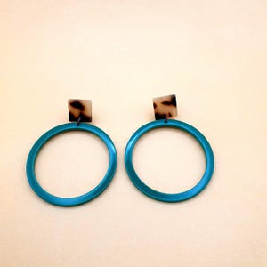 Può includere: Un paio di orecchini a cerchio turchese con borchie quadrate con motivo tartarugato. Gli orecchini hanno una finitura lucida e un design semplice e moderno. I cerchi sono di un vibrante colore turchese, e le borchie sono marroni e beige.
