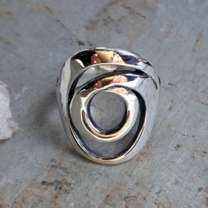 May include: A silver ring with a unique, abstract design featuring two overlapping circles. The ring is polished and has a smooth, shiny finish.