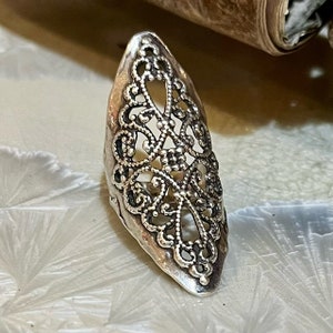 Puede incluir: Un anillo de filigrana plateado con forma ovalada puntiaguda. El anillo está hecho de diseños intrincados y delicados y tiene una estética vintage.