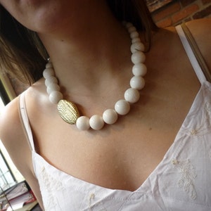 Puede incluir: Un collar de cuentas blancas con un colgante ovalado dorado. El collar lo lleva una persona con una blusa blanca.