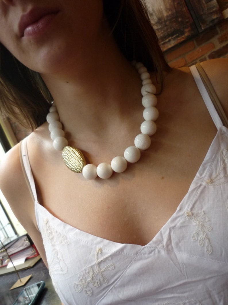Puede incluir: Un collar de cuentas blancas con un colgante ovalado dorado. El collar lo lleva una persona con una blusa blanca.