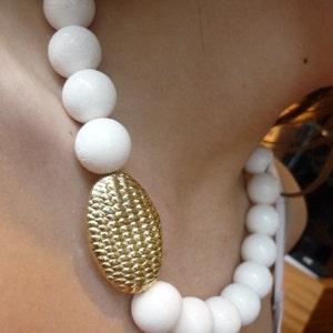 Puede incluir: Un collar de cuentas blancas con un colgante ovalado de oro. Las cuentas son grandes y redondas.