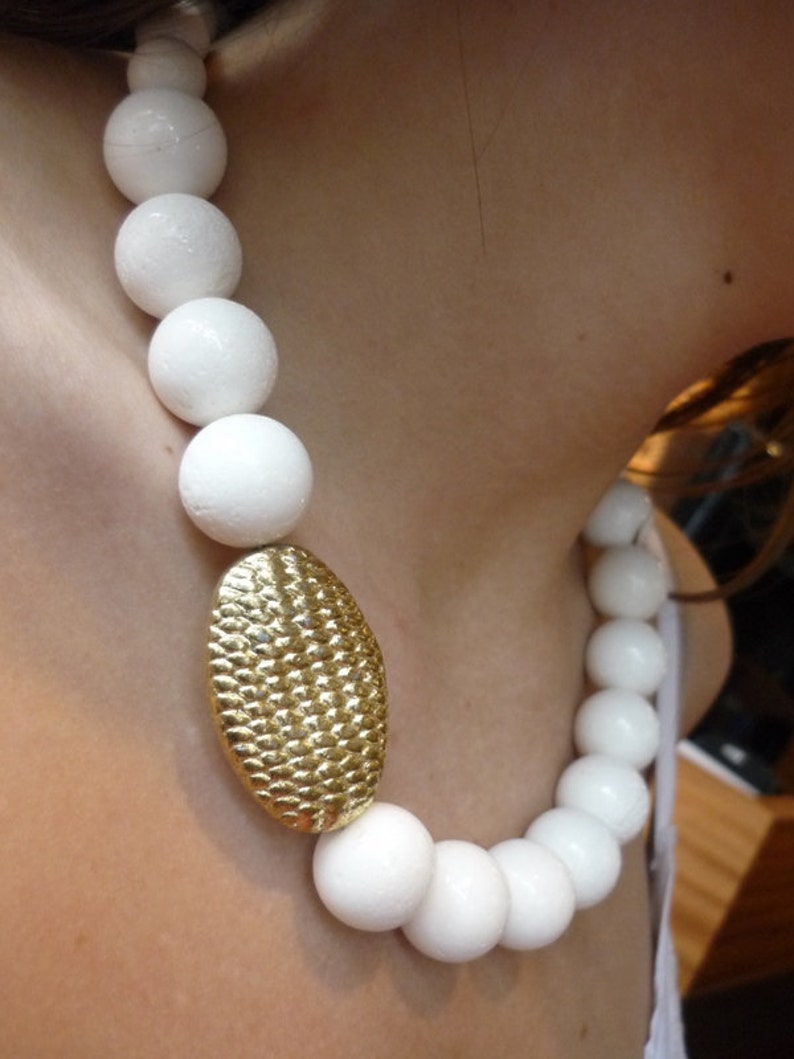 Puede incluir: Un collar de cuentas blancas con un colgante ovalado de oro. Las cuentas son grandes y redondas.
