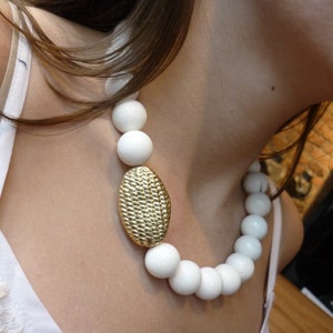 Puede incluir: Un collar de cer&aacute;mica blanca con cuentas grandes y redondas y un colgante ovalado dorado. El collar lo lleva una persona con el pelo largo y casta&ntilde;o.