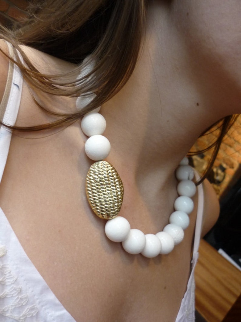 Puede incluir: Un collar de cer&aacute;mica blanca con cuentas grandes y redondas y un colgante ovalado dorado. El collar lo lleva una persona con el pelo largo y casta&ntilde;o.
