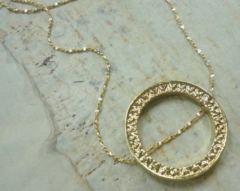 May include: A gold necklace with a delicate chain and a large, round pendant. The pendant is intricately detailed with a textured, ornate design.