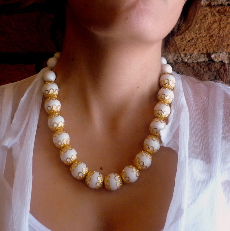 May include: A white beaded necklace with gold accents. The beads are round and have a delicate, intricate design. The necklace is worn by a person with a white blouse.