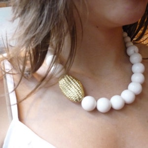 Puede incluir: Un collar de cuentas blancas con un colgante ovalado dorado. El collar est&aacute; hecho de cuentas grandes y redondas y lo lleva una persona con el pelo largo y casta&ntilde;o.