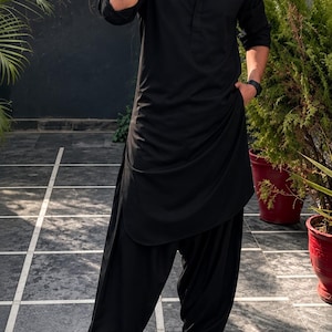 Könnte beinhalten: Ein einfarbig schwarzes, langärmeliges Kurta-Hemd mit spitzem Kragen und eine dazu passende, locker sitzende, gefaltete Shalwar-Hose. Das Outfit wird mit schwarzen Leder-Loafern und dunkel getönten Sonnenbrillen für einen Look kombiniert.