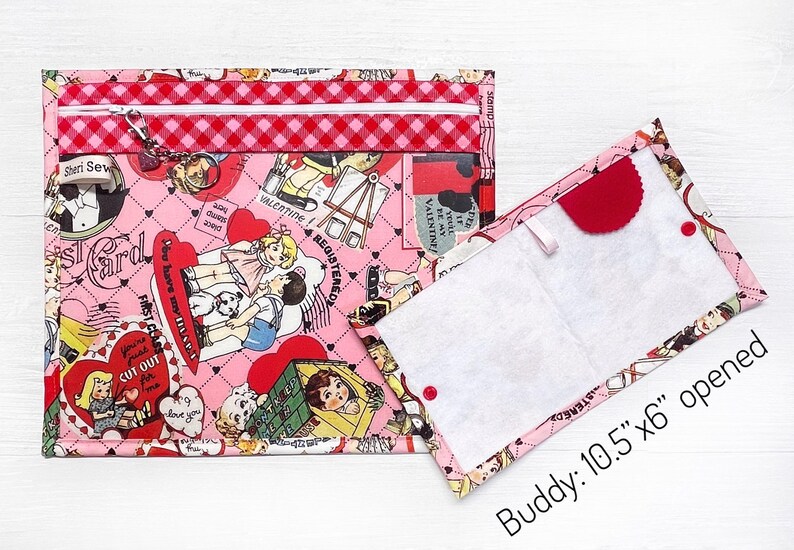May include: A pink zippered pouch with a quilted pattern and a white zipper. The pouch is decorated with a Valentine's Day theme print featuring hearts, flowers, and children. The pouch is open to reveal a white fabric lining. The text "Buddy: 10.5"x6" opened" is printed below the pouch.
