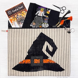 May include: A black and white striped fabric zippered pouch with a black witch hat design. The pouch has a black and orange trim and a small black heart with the word "Sweet" on it. The pouch is open and a cross stitch pattern of a witch with a black cat and a pumpkin is visible inside.
