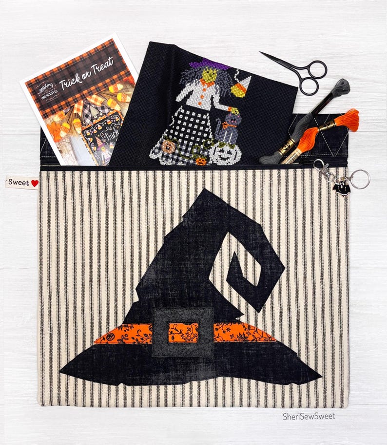 May include: A black and white striped fabric zippered pouch with a black witch hat design. The pouch has a black and orange trim and a small black heart with the word "Sweet" on it. The pouch is open and a cross stitch pattern of a witch with a black cat and a pumpkin is visible inside.