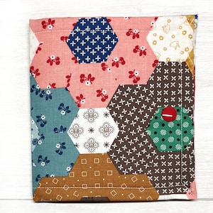 May include: A patchwork fabric square with a hexagonal pattern in shades of pink, blue, brown, green, and white. The fabric has a variety of small patterns, including polka dots, floral prints, and geometric designs. A red button is sewn onto the fabric.