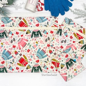 May include: Three zippered pouches with a white background and a repeating pattern of colorful sweaters, mugs, and snowflakes. The pouches are made of fabric and have a zipper closure. The text "SheriSewSweet" is printed on the bottom of the largest pouch.