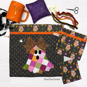 May include: Three zippered pouches in black fabric with a ghost and autumn leaf pattern, featuring an orange zipper and a large ghost applique with a leopard print bow. Includes an orange mug with the text SPOOKY, a purple pincushion, and embroidery floss.