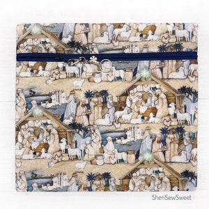 May include: A zippered pouch with a blue zipper and a snowflake charm. The pouch features a repeating pattern of a nativity scene with a stable, animals, and people. The text "SheriSewSweet" is printed on the bottom of the pouch.