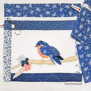 May include: A blue and white quilted zippered pouch with a bird applique. The pouch features a floral pattern, a white zipper, and a key ring with a flower charm. The bird is blue and orange, perched on a branch.
