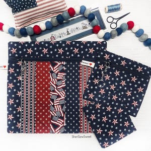 May include: A patriotic-themed set of zippered pouches and accessories. The pouches feature a navy blue star pattern, red and white stripes, and American flag accents. Additional items include a felt ball garland, scissors, and a spool of thread.